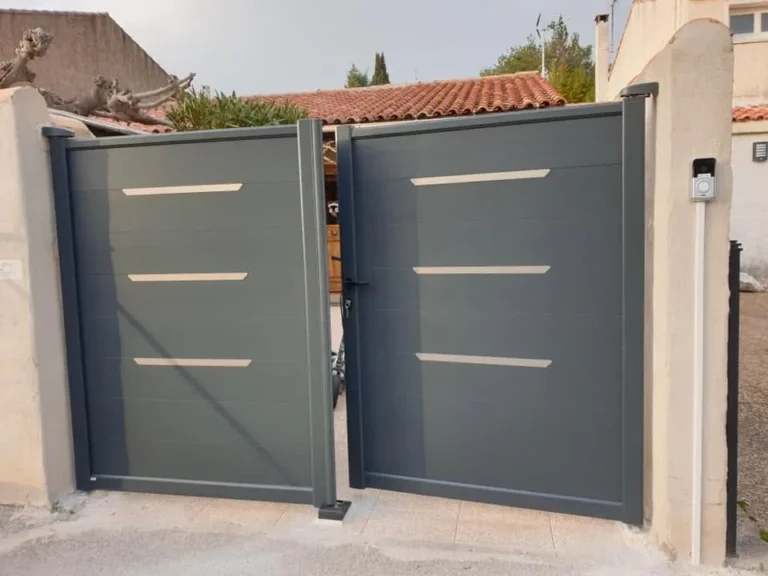 Portail battant en aluminium gris avec inserts inox devant une haie